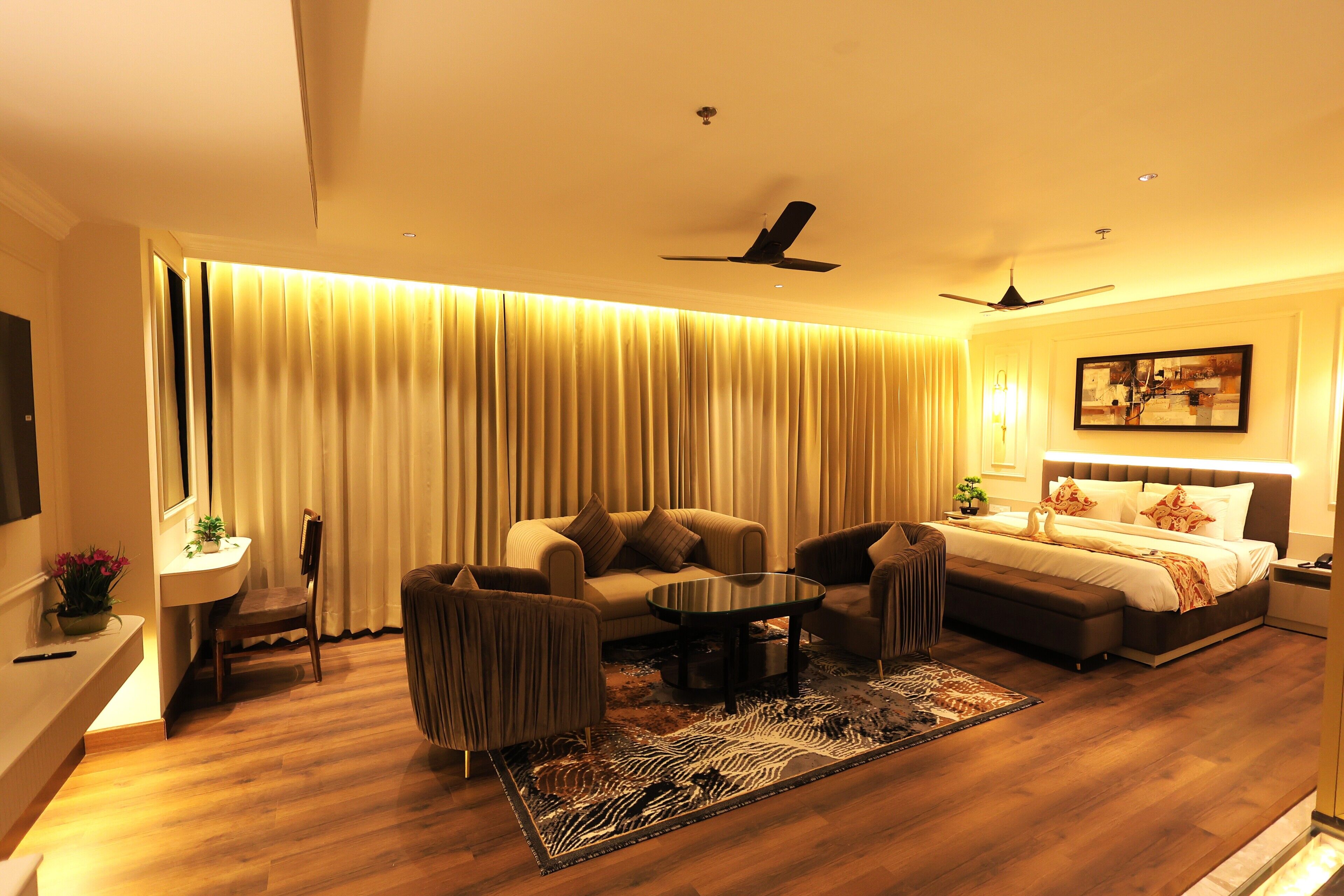 A modern hotel room with a sofa, armchair, and a bed with a headboard. There is a small table and a chair, a ceiling fan, and a large window with curtains.