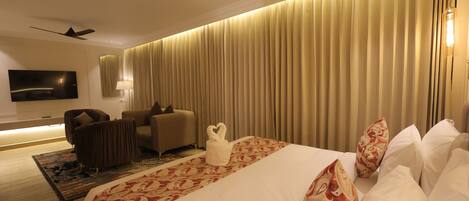 A hotel room with a large bed, a flat-screen TV mounted on the wall, a ceiling fan, and a large window with curtains.