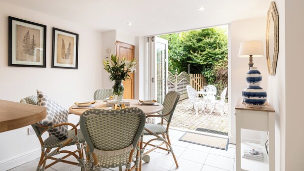 A dining area with wicker chairs, a wooden table, and a vase of flowers. There is a door leading to an outdoor patio with white chairs and a garden. - Two Cottages near Stratford-upon-Avon (Clifford Chambers)