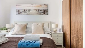 A neatly made bed with a brown quilt, two pillows, and a blue towel with "Quay Holidays" text. There is a wooden door to the right and a white nightstand with a lamp and a small plant. - Knots - Apartment close to Poole Quay, Poole Town (Poole)