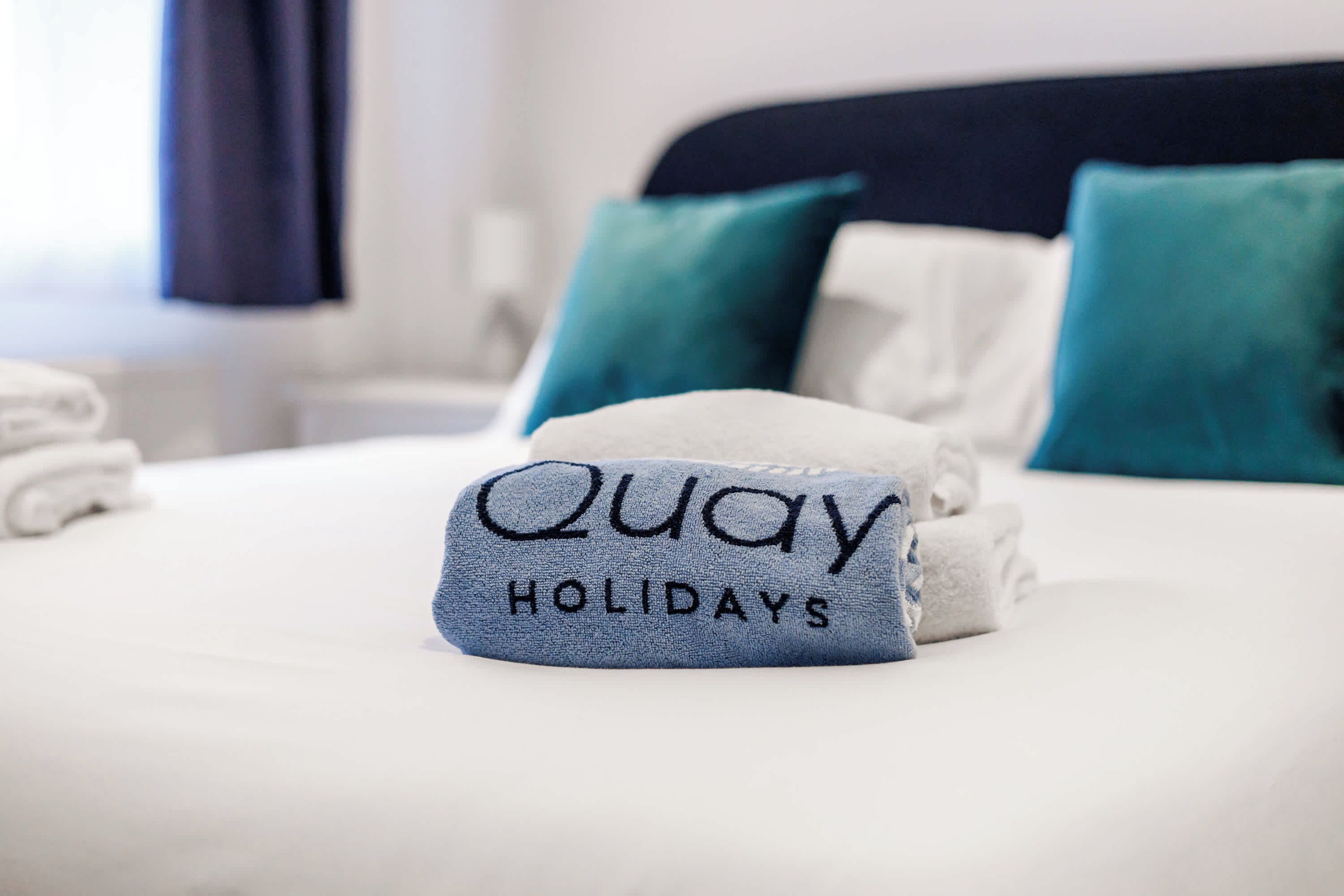 Una cama bien tendida con una toalla que dice 'Away HOLIDAYS' y dos almohadas color turquesa.