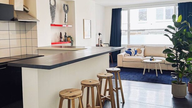 Et moderne køkken med en central ø, barstole, et spiseområde med sofa og bord, samt et stort vindue med gardiner.
