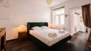 Een net opgemaakt bed met witte lakens en opgevouwen handdoeken, een bedlampje, een raam met gordijnen en een radiator.