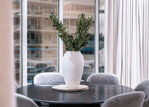 Ein modernes Zimmer mit einem runden Tisch, grau gepolsterten Stühlen und einer weißen Vase mit Grünpflanzen.
