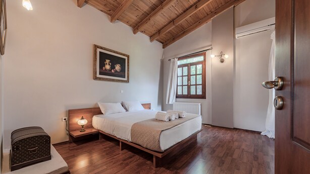 A bedroom with a wooden bed, a nightstand with a lamp, a painting on the wall, a window with curtains, and a door. - Skine Dream Villa, Charming Villa with Private Pool (Platanias)