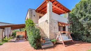 A rustic stone building with a wooden staircase leading to a porch, a swing chair, and a patio area with a table and chairs. - Skine Dream Villa, Charming Villa with Private Pool (Platanias)