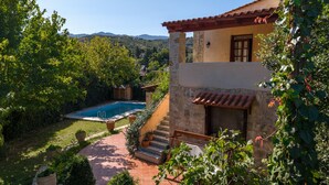 Una casa de dos pisos con piscina, rodeada de abundante vegetación y colinas al fondo.