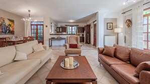 Amplia sala de estar con un sofá beige, un sofá de cuero marrón y una mesa de centro de madera. Hay un comedor con mesa y sillas, y una cocina al fondo.