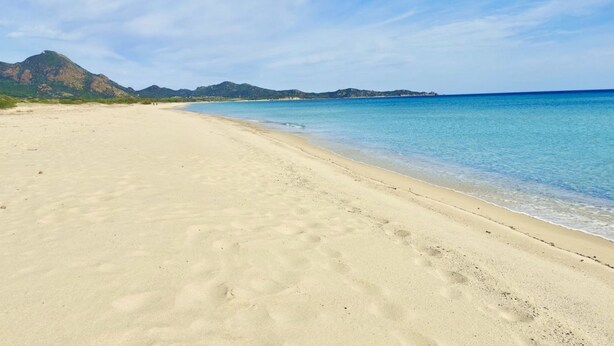 Een breed zandstrand met helderblauw water en op de achtergrond bergen.