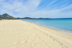 Ein weiter, sandiger Strand mit klarem, blauem Wasser und Bergen im Hintergrund.