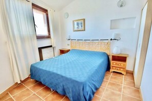 A bedroom with a bed, a nightstand, a lamp, a window with curtains, and a wall clock. - Rosemary Villa 6 beds (Muravera)