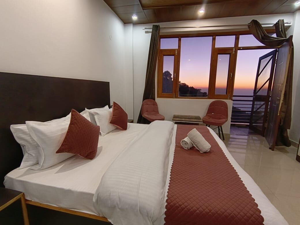 Habitación de hotel con una cama, vistas al atardecer a través de amplios ventanales y un balcón con sillas.