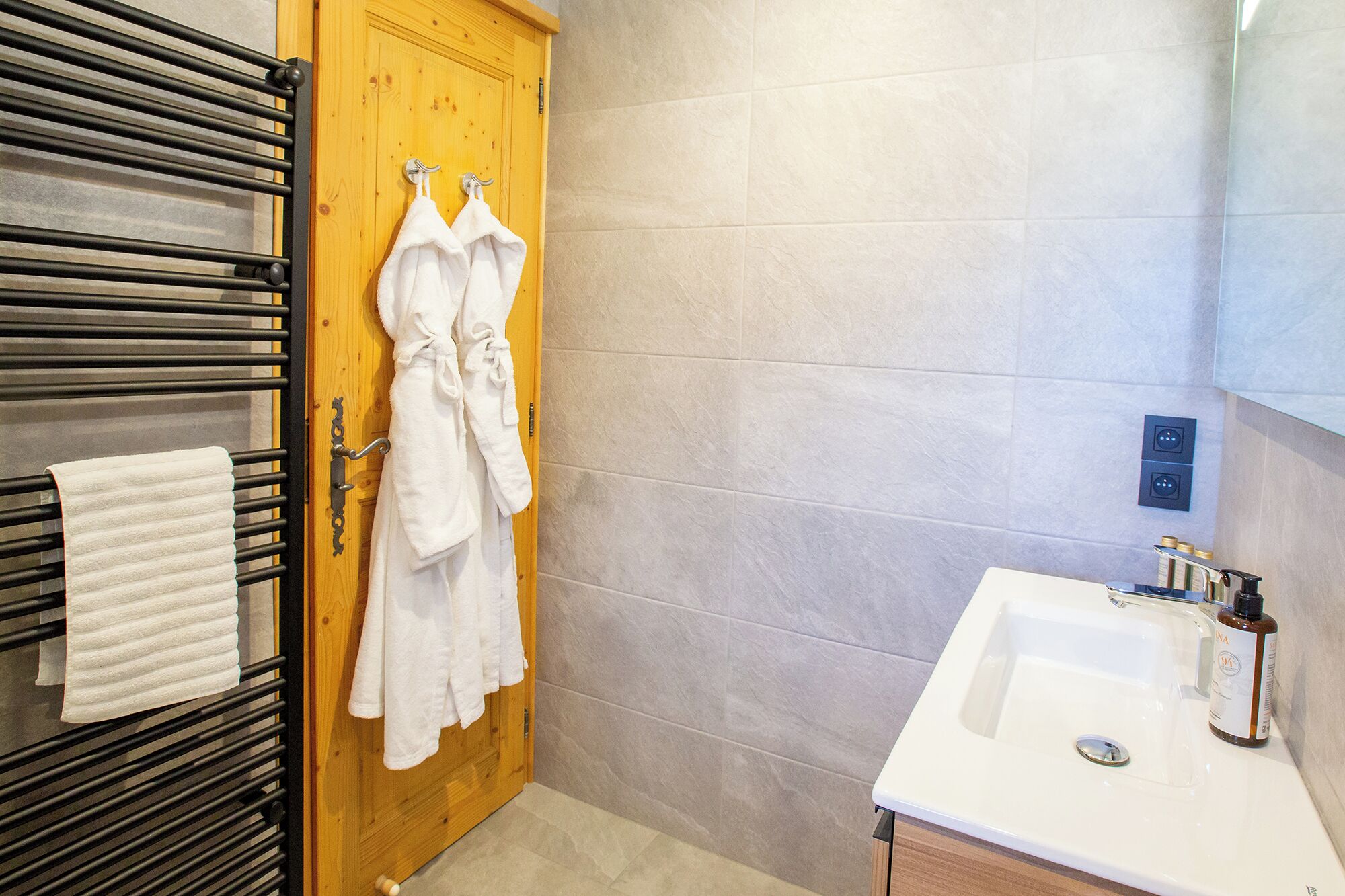 Um banheiro com porta de madeira, porta-toalhas com toalhas brancas, pia com um frasco e parede revestida de azulejos.