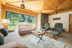 A cozy living room with a stone fireplace, a large window offering a view of greenery, and a comfortable seating area with a sofa and two armchairs.