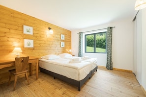 A room with a wooden wall, a bed with white bedding, a desk with a chair, and a window with curtains.