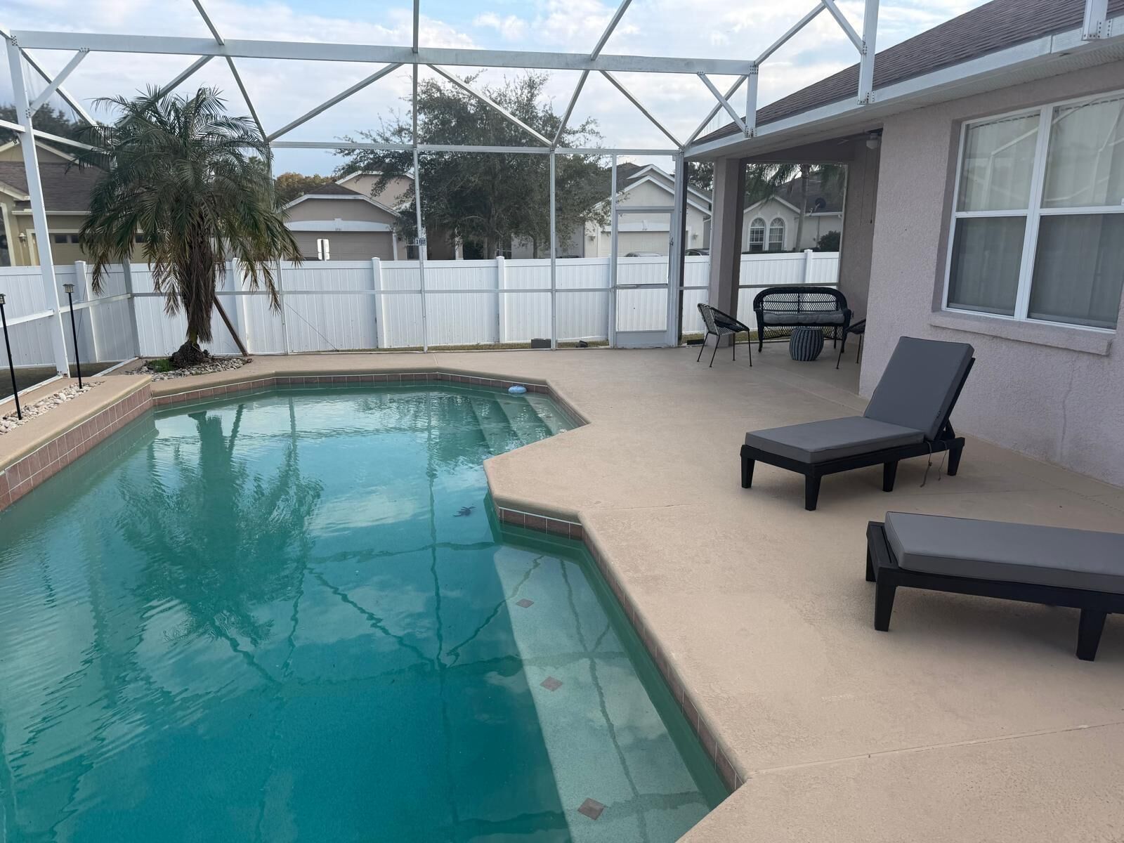 A swimming pool with a white fence, lounge chairs, and a covered patio area.