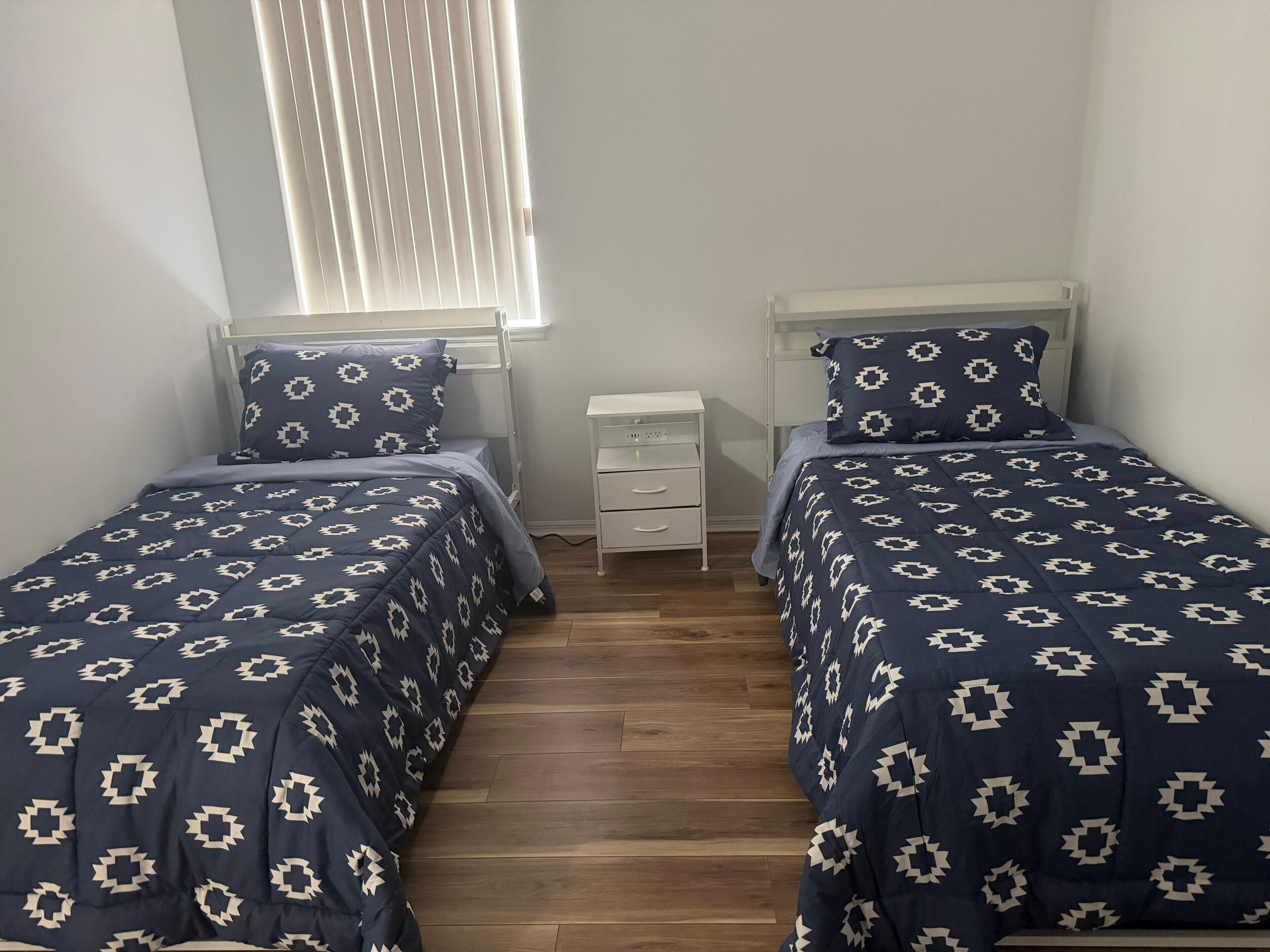 Un dormitorio con dos camas individuales, cada una con una colcha estampada y almohadas a juego, una mesita de noche con cajones y una ventana con persianas.