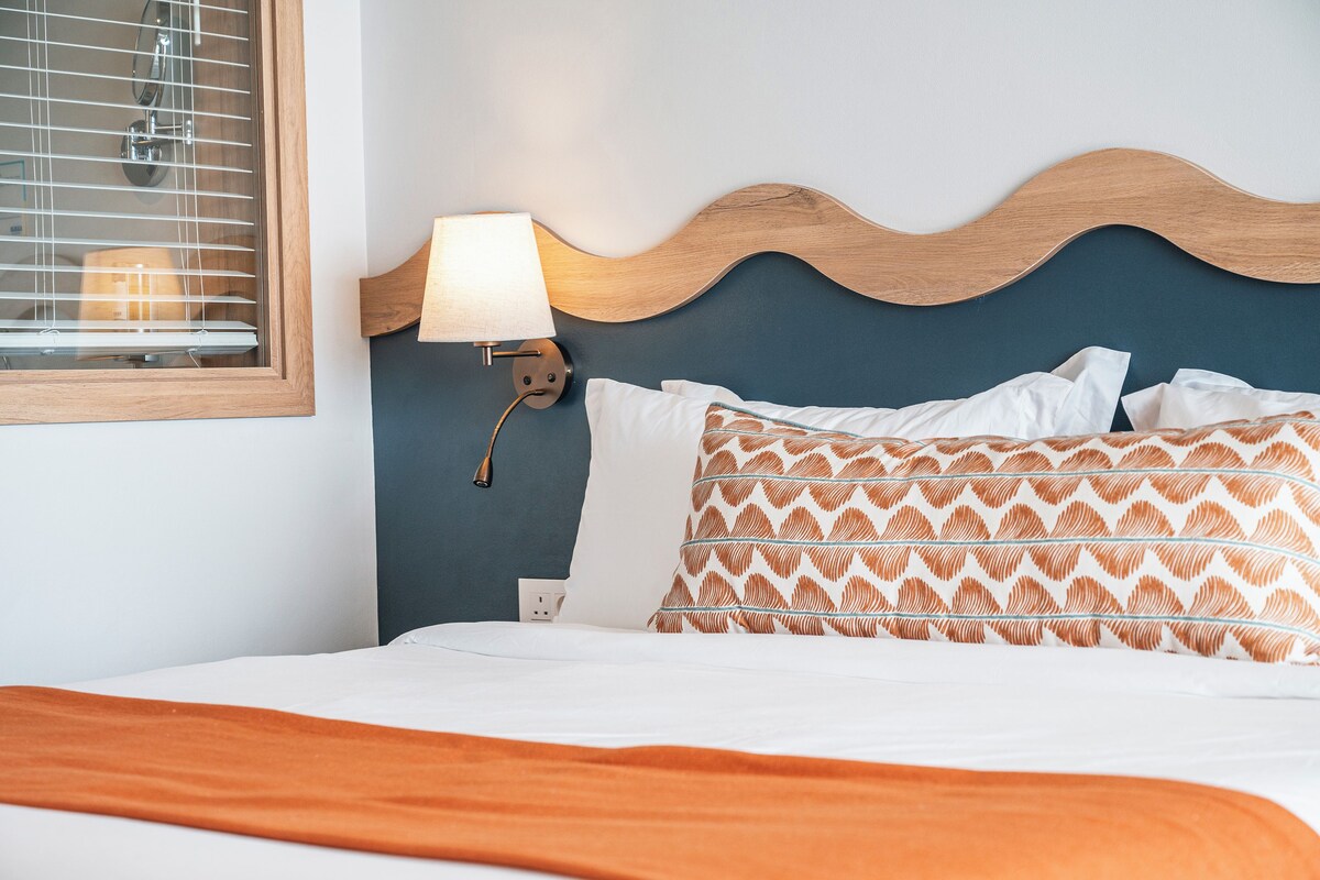 A neatly made bed with white and patterned pillows, a wooden headboard, a wall-mounted lamp, and a window with blinds.