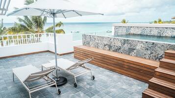 A balcony with a view of the ocean, a lounge chair, a small table, and a cart.