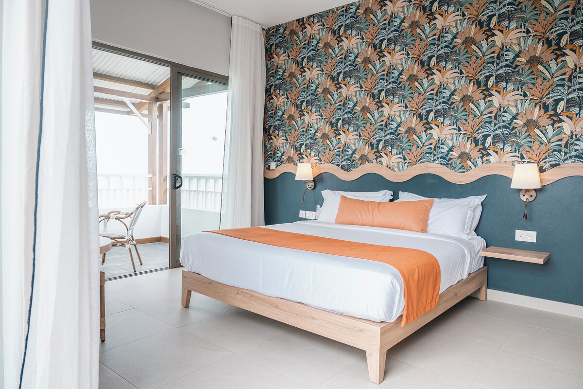 A modern bedroom with a wooden bed, white and orange bedding, a floral wallpaper, and a balcony with a chair.