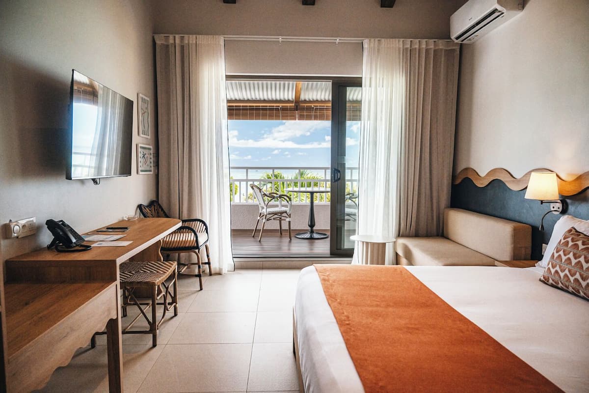 Ein Hotelzimmer mit Bett, Schreibtisch mit Telefon, Stuhl, Sofa, Balkon mit Aussicht und Klimaanlage.