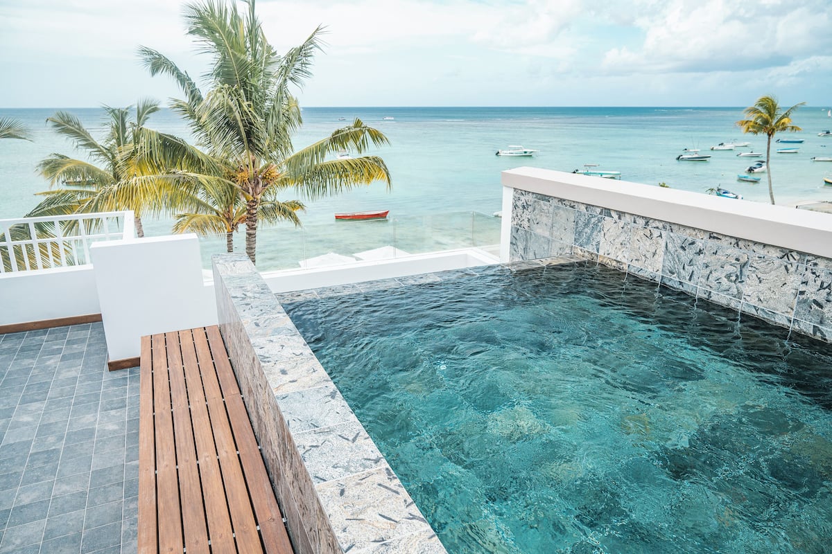 Blick auf einen Pool mit Holzterrasse und Steinmauer, mit Blick auf ein ruhiges Meer mit Booten und Palmen.