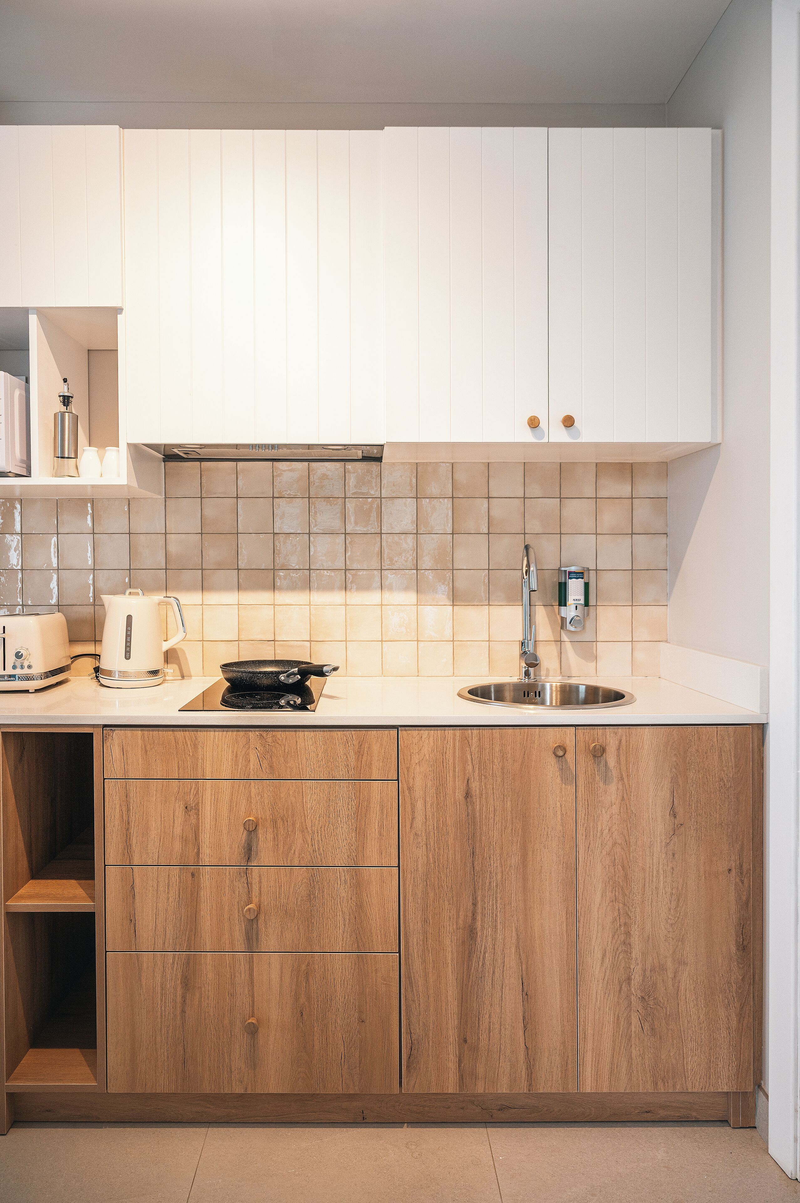 Une cuisine moderne avec des meubles blancs, un pare-feu en carrelage, un évier en acier inoxydable, une bouilloire, un grille-pain et des meubles bas en bois.