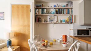 Een eetruimte met een ronde tafel, witte stoelen, ontbijtartikelen en een boekenplank met boeken en decoratieve objecten.