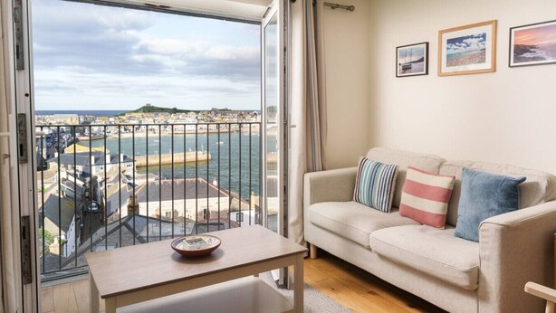 Een balkon met uitzicht op een haven en gebouwen, een bank met kussens en een klein tafeltje met een schaal.