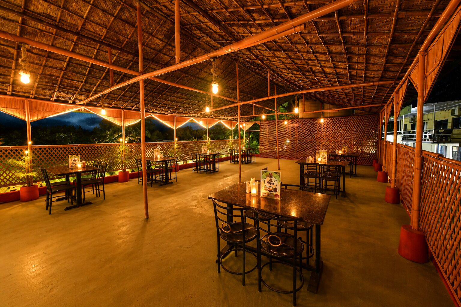 An outdoor dining area with tables and chairs, a thatched roof, and string lights.