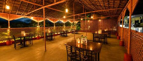 An outdoor dining area with tables and chairs, a thatched roof, and string lights.
