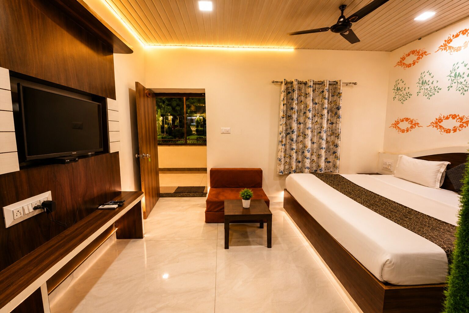 A hotel room with a bed, a TV, a sofa, a small table with a plant, and a ceiling fan.