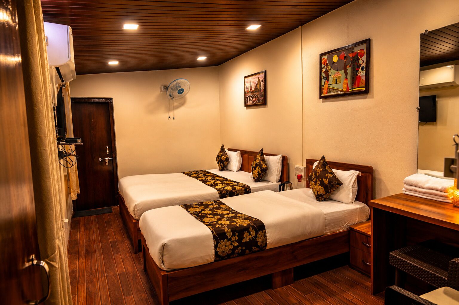 A hotel room with two beds, a wooden ceiling, a wall-mounted fan, a painting, and a wooden desk with a mirror.