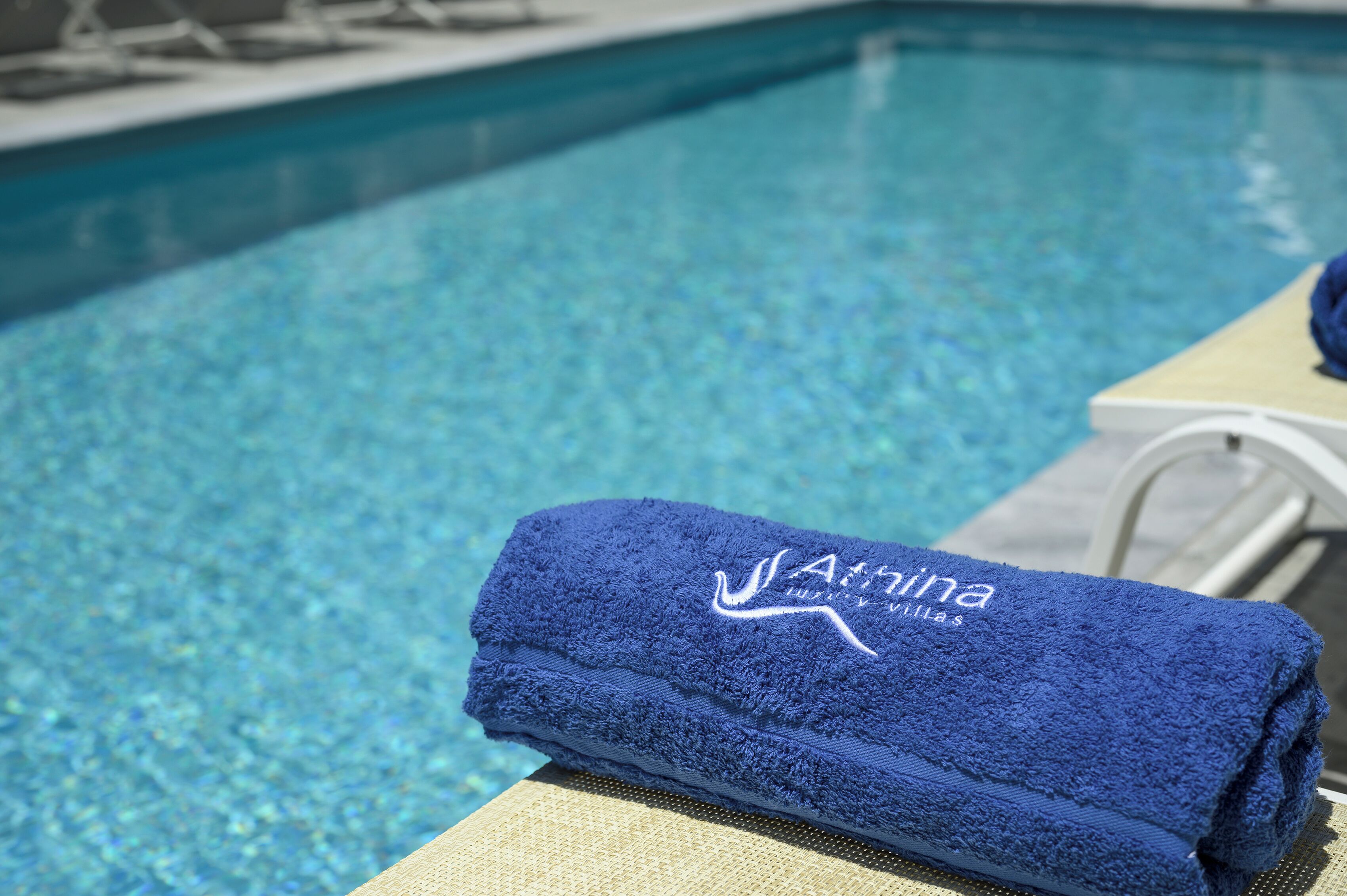 A poolside area with a blue towel labeled "Athina Luxury Villas" on a lounge chair.