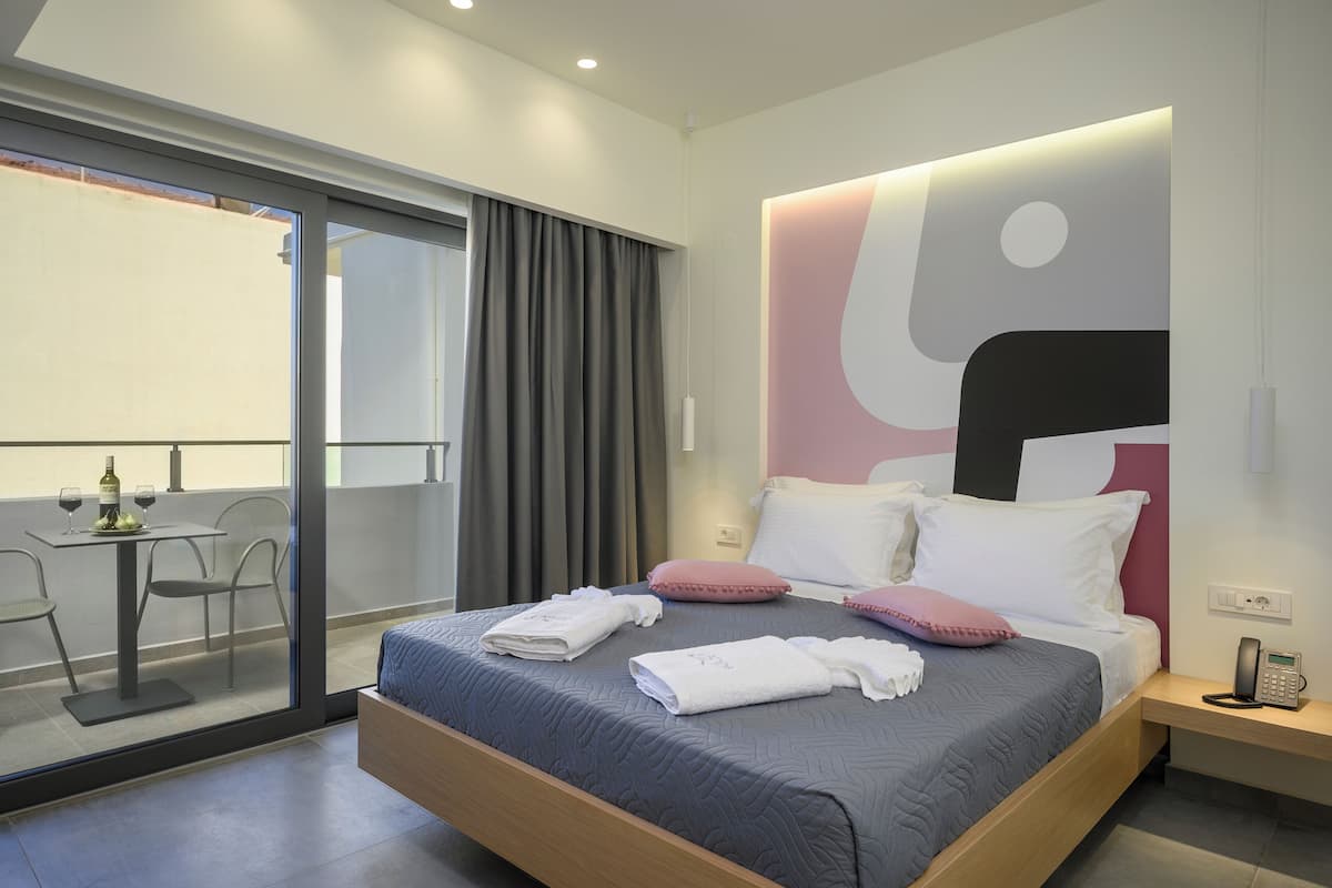 Ett modernt hotellrum med en stor säng, ett nattduksbord med en telefon och utsikt genom en glasdörr till en balkong med ett bord och stolar.