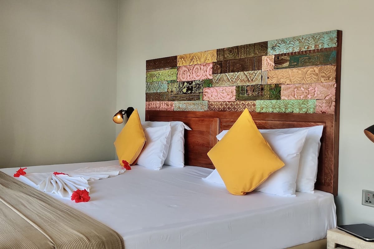 A neatly made bed with white linens and yellow pillows, a wooden headboard with a decorative panel, and a bedside table with a lamp and a tablet.
