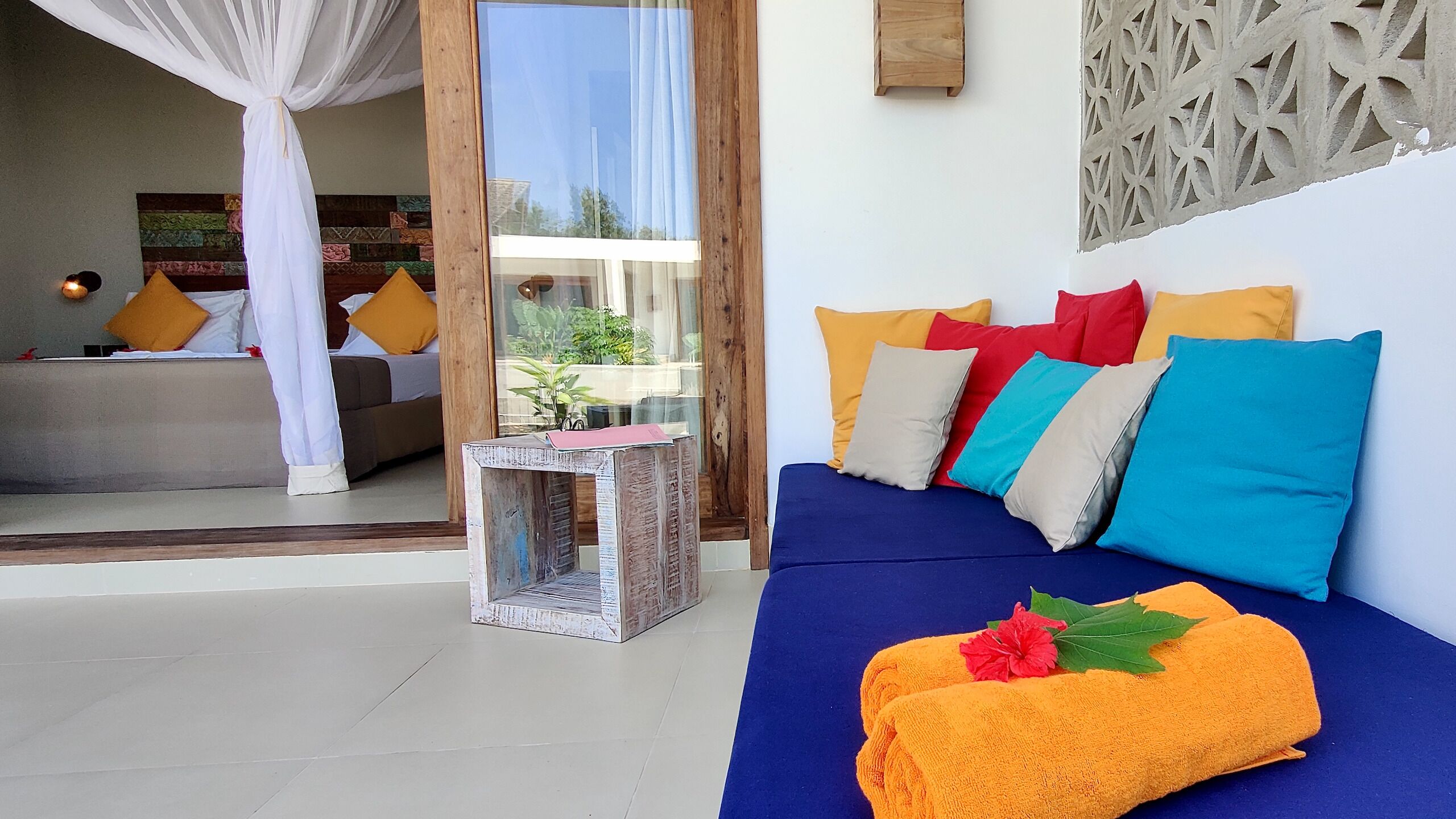 Una habitación con un sofá azul, cojines de colores, una mesita con una planta y una cortina blanca.