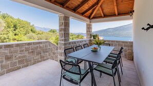 A covered outdoor patio with a stone wall, a table with chairs, and a view of a mountainous landscape. - Luxury 4-bedroom Villa with Private Heated Pool & Sea View, near Portonovi. ( Đenovići)