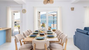 A dining area with a wooden table set for six, surrounded by beige chairs, with a view of the sea and mountains through large windows. - Luxury 4-bedroom Villa with Private Heated Pool & Sea View, near Portonovi. (Đenovići)
