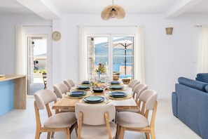 A dining area with a wooden table set for six, surrounded by beige chairs, with a view of the sea and mountains through large windows. - Luxury 4-bedroom Villa with Private Heated Pool & Sea View, near Portonovi. (Đenovići)