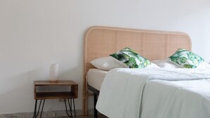 Una cama bien hecha con ropa blanca y dos almohadones decorativos, una mesita de noche de madera con un florero blanco, y una pared lisa.