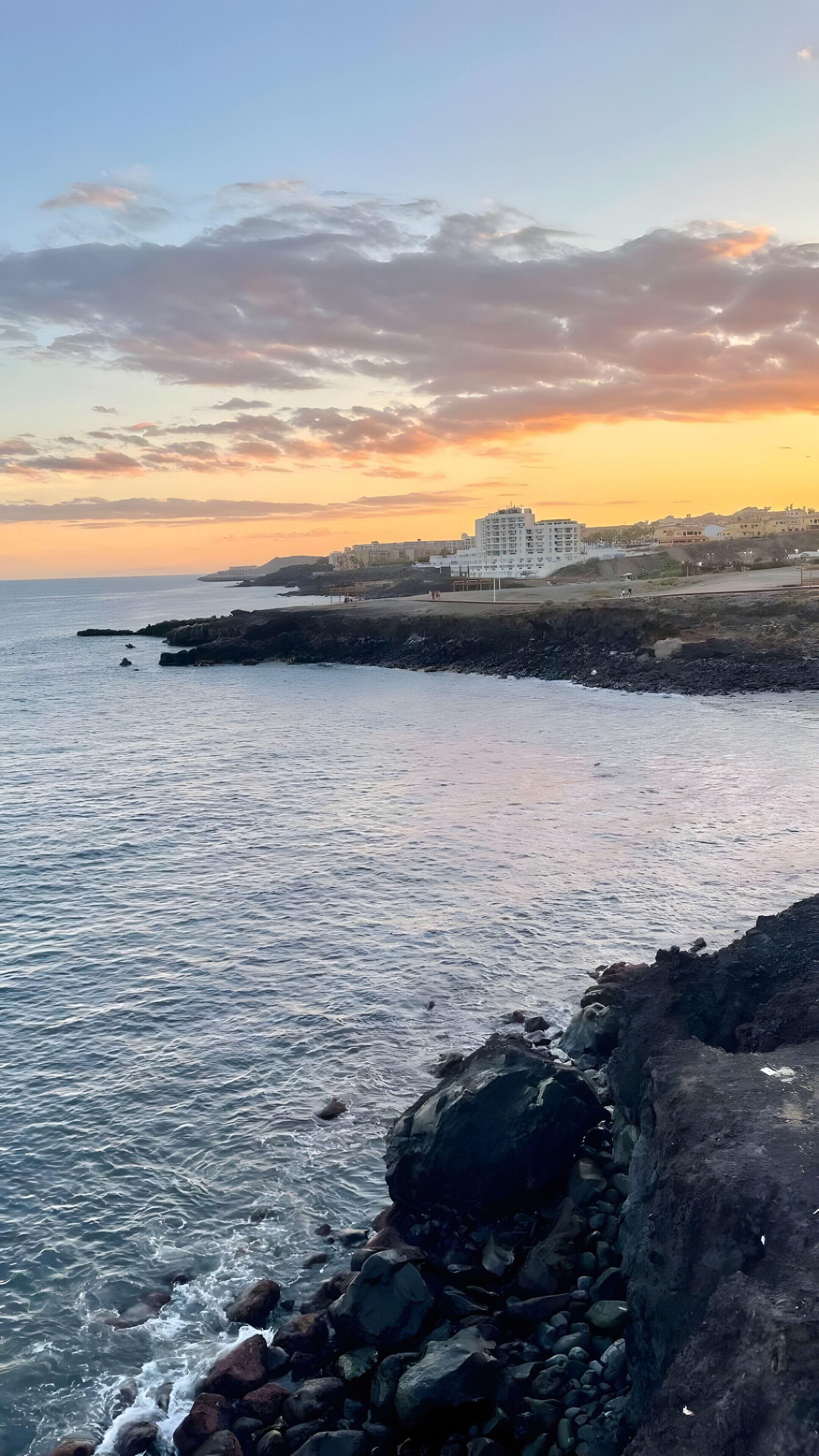 A coastal view with a rocky shoreline, a building with multiple floors, and a sunset sky.