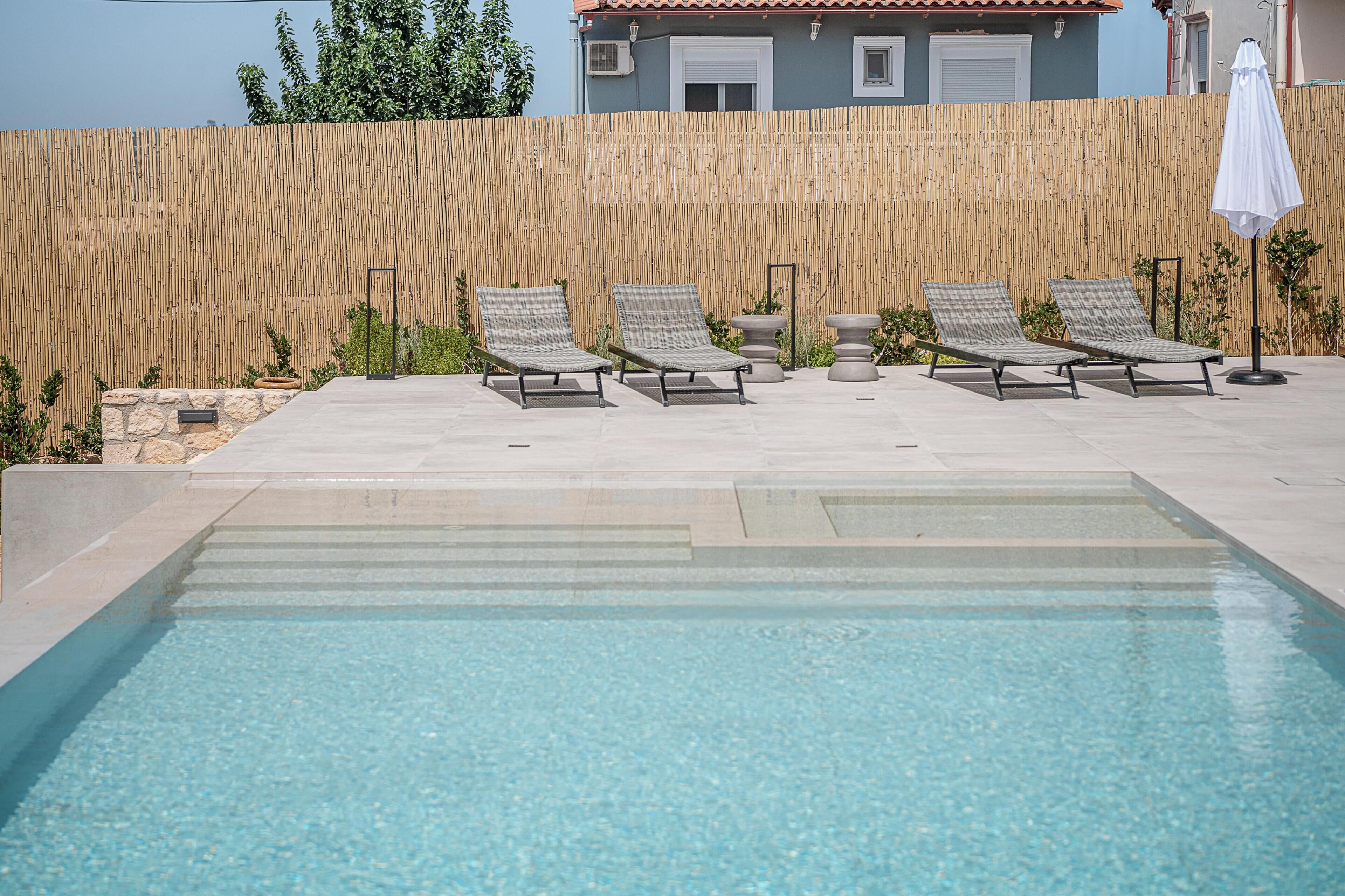 Une piscine entourée d’une clôture en bambou et d’un mur de pierre, avec des chaises longues et un parasol.