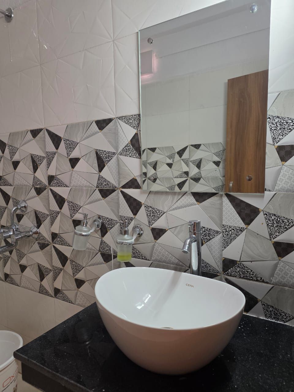 Une salle de bain avec un lavabo blanc, un plan de toilette noir et un miroir au-dessus. Les murs sont recouverts de carreaux disposés selon un motif géométrique.