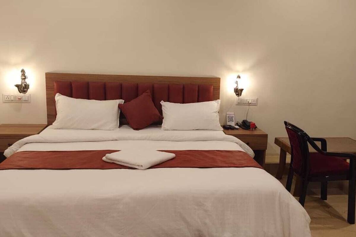 Quarto de hotel com uma cama, duas mesas de cabeceira, uma cadeira, uma mesinha e uma cortina.