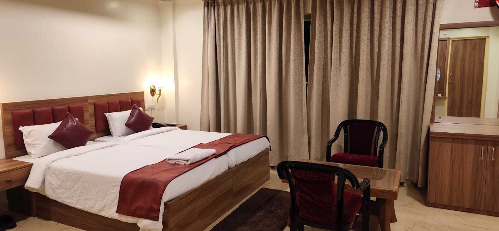 Ett hotellrum med en säng i trä, ett skrivbord, en stol, en spegel och ett fönster med gardiner.