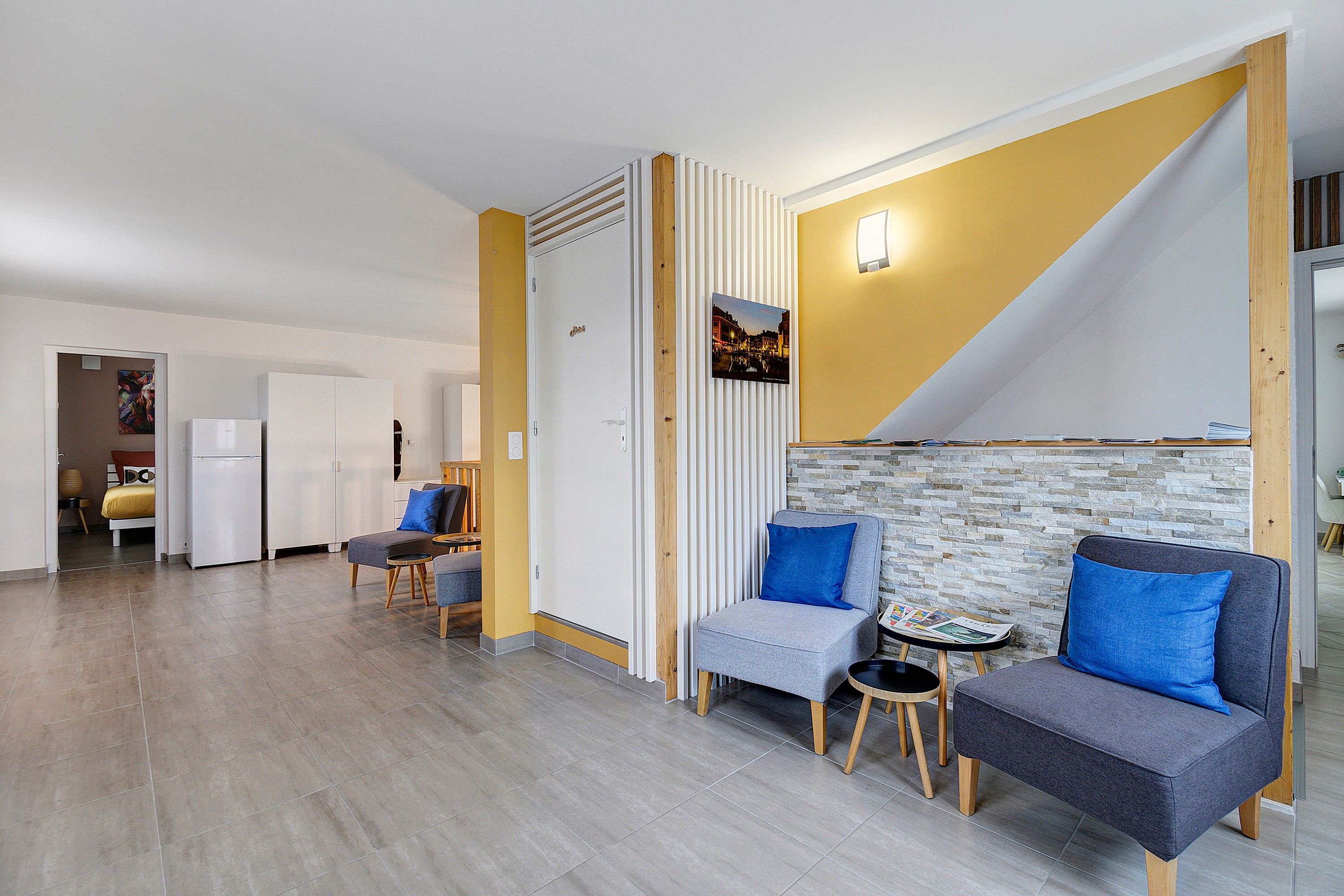 Een moderne woonkamer met een bank, fauteuils en een bijzettafeltje. Achtergrond is een keuken met een koelkast en een eettafel.