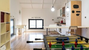 A room with a foosball table, a desk, and a chair. There is a window with blinds, a shelf with storage, and a lamp. - La Manovella Apartment (Caselle Torinese)
