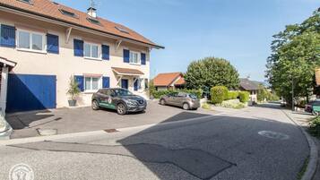 Une rue résidentielle avec deux voitures garées, une maison aux volets bleus et un ciel dégagé.