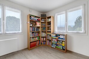 Een hoekappartement met een grote boekenkast vol boeken en spelletjes, twee ramen met uitzicht op bomen en een radiator aan de muur.